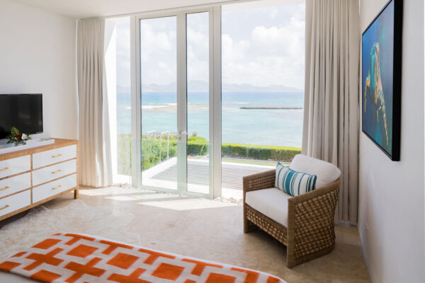 Blue Queen Suite at Champagne Shores Villa, featuring a large window with ocean views, a white dresser with gold handles, a comfortable chair with a striped cushion, and a bed with an orange patterned blanket.