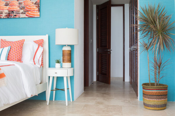 Blue Queen Suite at Champagne Shores Villa, showcasing a white bed with vibrant orange and pink pillows, a white nightstand with a rustic lamp and a small succulent, against a turquoise accent wall. An open door leads to the hallway, with a decorative plant in a colorful woven basket nearby.