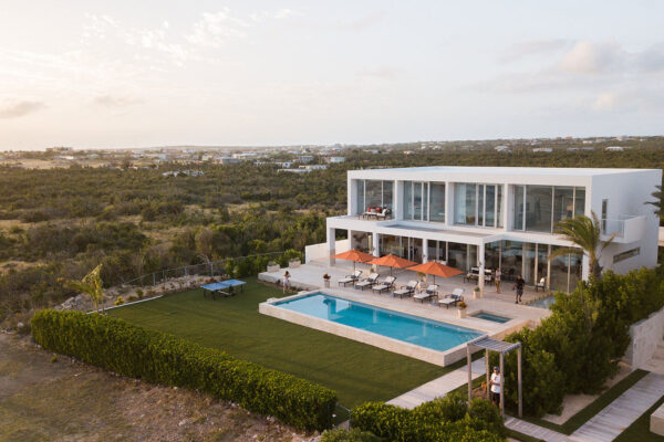 Aerial view of Champagne Shores Villa, showcasing its modern architecture with large windows, a spacious pool area with sun loungers and orange umbrellas, surrounded by lush greenery and scenic landscapes.