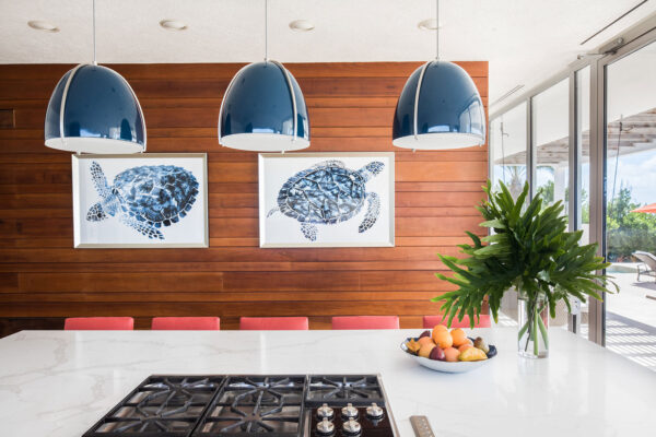 Modern kitchen at Champagne Shores Villa, featuring a white marble island with red cushioned bar stools, three blue pendant lights, and two framed turtle prints on a wooden accent wall.