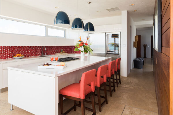 Fully-equipped European kitchen at Champagne Shores Villa, featuring a large white island with red upholstered barstools, modern pendant lights, and a sleek red backsplash. The kitchen includes stainless steel appliances, a flower arrangement, and a tray with drinks and snacks on the island.