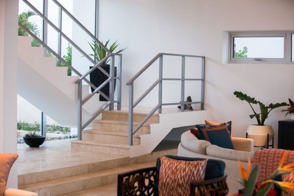 Interior view of Champagne Shores Villa, showcasing a modern staircase with light stone steps and minimalist metal railings, a cozy seating area with colorful cushions, and large windows letting in natural light.