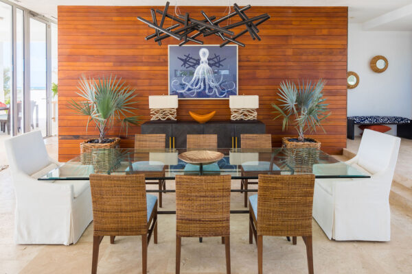 Dining area at Champagne Shores Villa, featuring a glass-top table with wicker and white upholstered chairs, a modern black chandelier, and a wooden accent wall with a framed octopus print. Two potted palm plants and decorative table lamps complete the stylish setting.