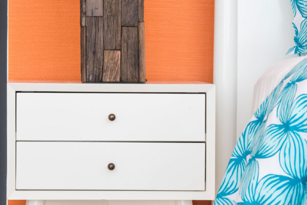 Close-up of a bedside table in orange queen suite at Champagne Shores Villa, featuring a white two-drawer nightstand with a rustic wooden lamp against a vibrant orange wall. The bed has a blue and white leaf-patterned duvet.