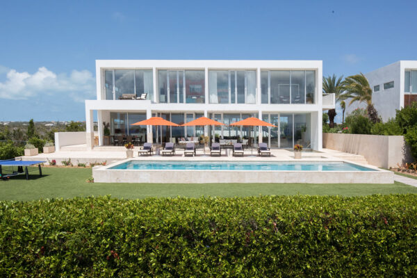 Exterior view of Champagne Shores Villa, featuring modern architecture with large windows, a pool surrounded by orange umbrellas and lounge chairs, a green lawn, and a clear blue sky.