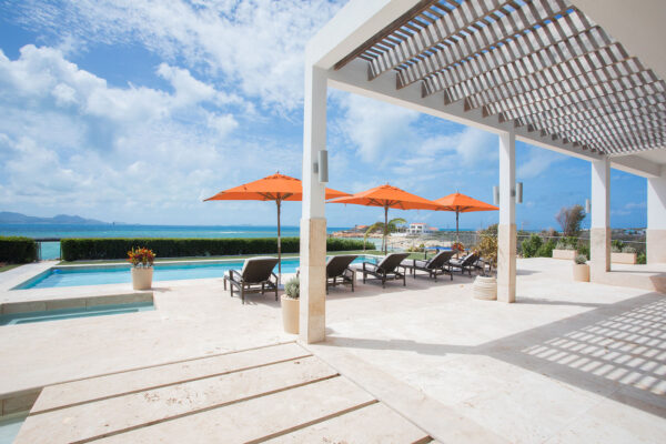 Oceanfront terrace at Champagne Shores Villa with sun loungers and orange umbrellas by the heated pool, overlooking the turquoise waters of Anguilla's Pelican Bay.