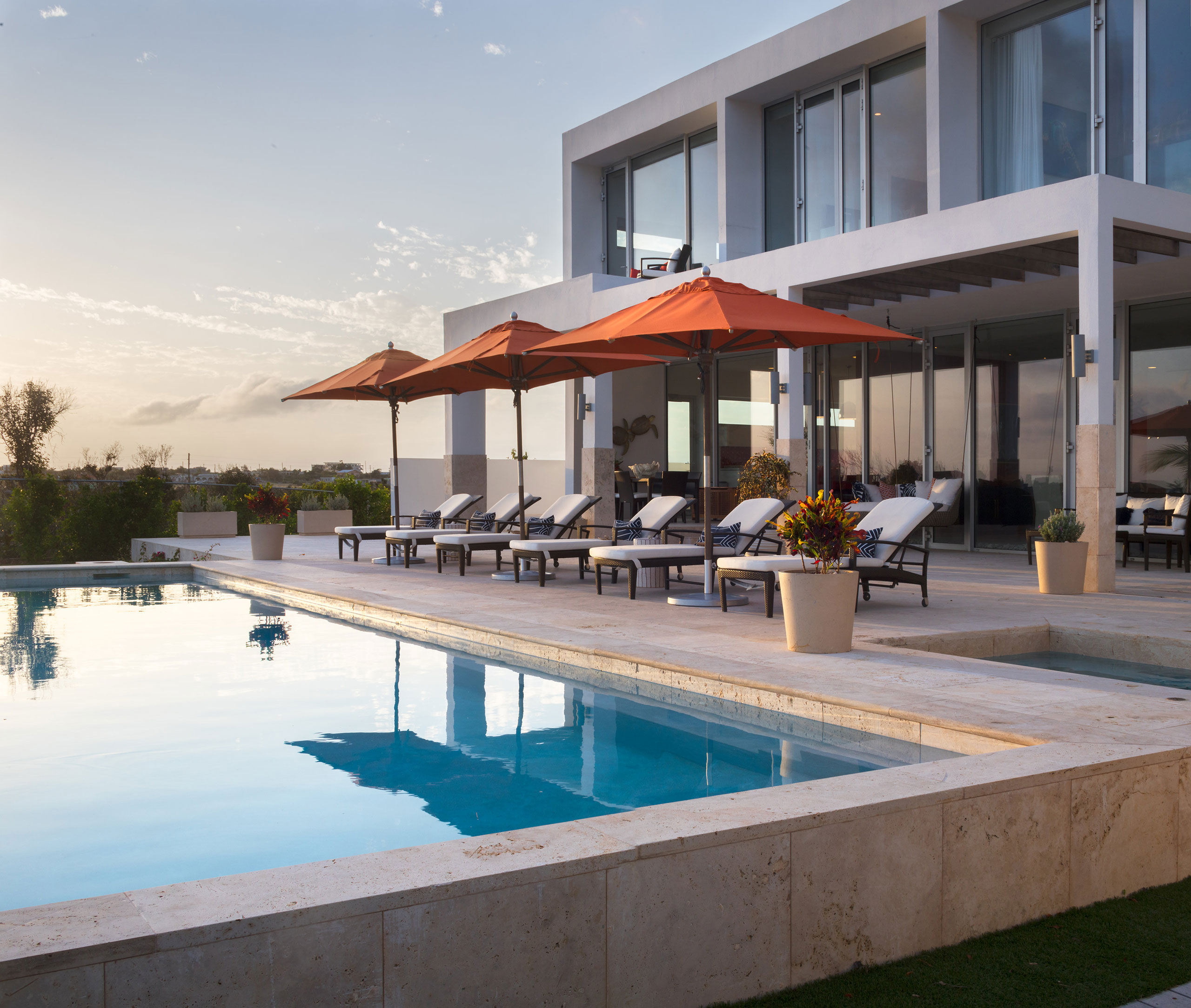 Luxurious poolside area at Champagne Shores Villa in Anguilla with sun loungers under orange umbrellas, adjacent to the modern villa and overlooking serene landscapes.