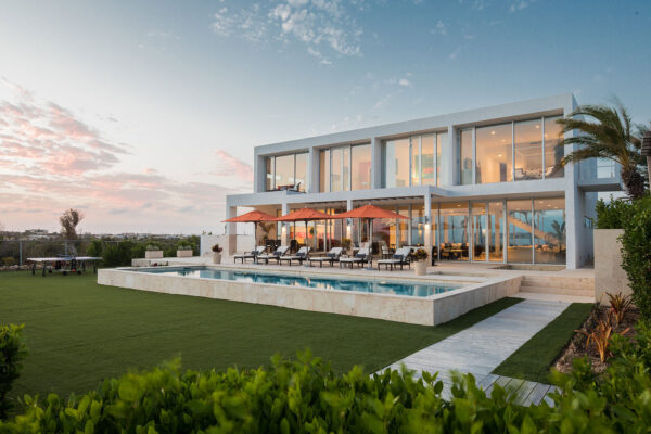 Exterior view of Champagne Shores Villa at sunset, showcasing the modern architecture, pool area with sun loungers and umbrellas, and a manicured lawn.