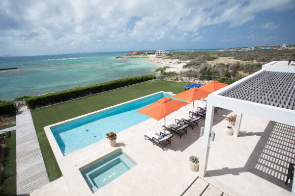 View of the heated pool and turquoise waters from the oceanfront terrace at Champagne Shores Villa in Anguilla, with sun loungers and orange umbrellas.