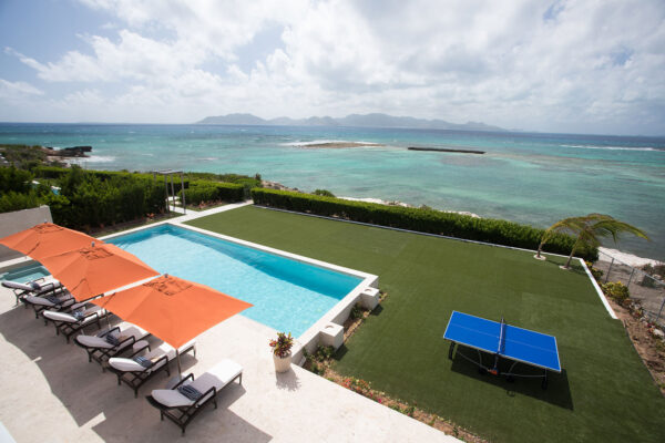 View from Champagne Shores Villa, showcasing a pool with orange umbrellas and lounge chairs, a lush green lawn with a ping pong table, and a stunning ocean backdrop with clear blue water St. Martin in the distance.