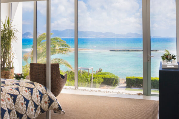 Secondary Primary Suite at Champagne Shores Villa in Anguilla with floor-to-ceiling windows showcasing a stunning ocean view, featuring a comfortable bed with patterned bedding and a wicker chair.