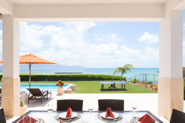 Outdoor dining area under terrace at Champagne Shores Villa, featuring a glass-top table set with red napkins and white plates, overlooking the pool, lawn, and ocean with a stunning view of the horizon.