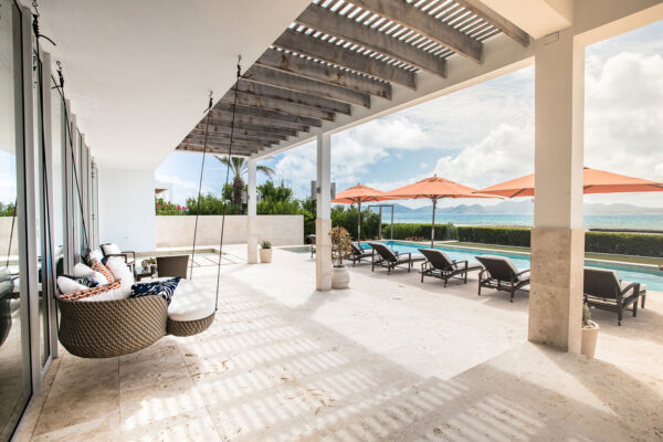 Terrace at Champagne Shores Villa, featuring cozy hanging chairs with blue and white pillows, a pergola providing partial shade, and a view of the pool with orange umbrellas, lounge chairs, and the ocean in the background.